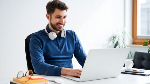 Ein junger Mann tippt zu Hause fröhlich an einem Notebook.