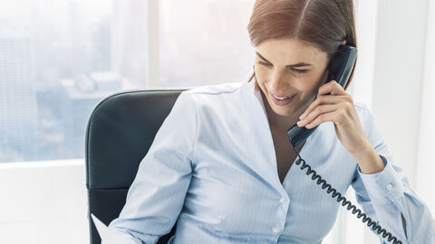 Eine Frau sitzt im Büro. Sie hält in einer Hand ihre Unterlagen und telefoniert gleichzeitig.